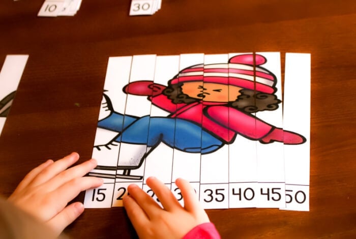 horizontal image with photo of a child's hands putting together a line up skip counting puzzle with the picture of a girl in winter hat, gloves and ice skates with skip counting numbers by 5s.