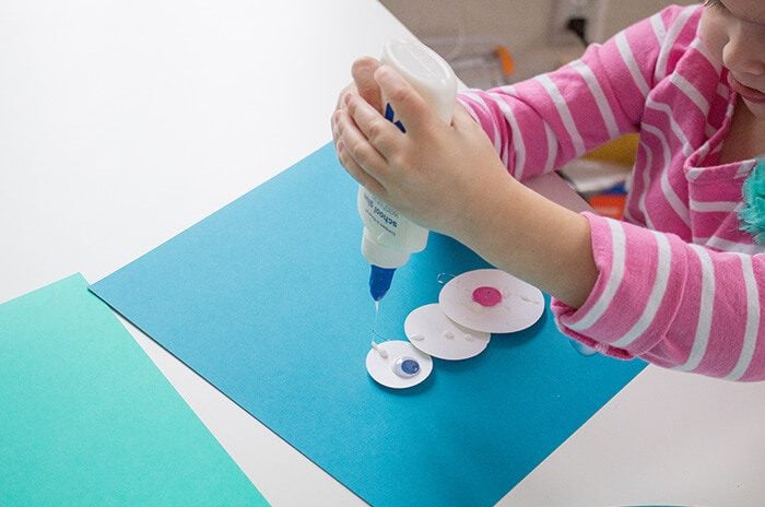 a snowman made of three white paper circles is on a piece of blue construction paper, and a child is adding googly eyes.