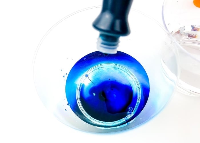 How to Dye Seashells for a Summer Sensory Bin 10 dropping blue food coloring from the bottle into water in a clear cup.