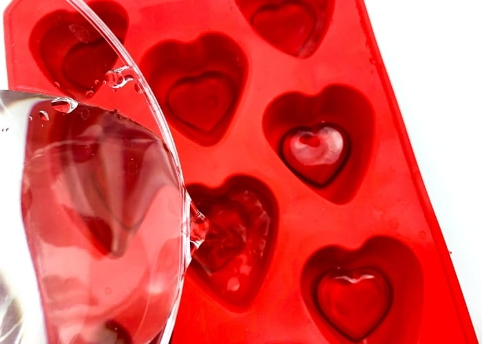 pouring water into a heart shaped ice cube tray with red heart gems in them.