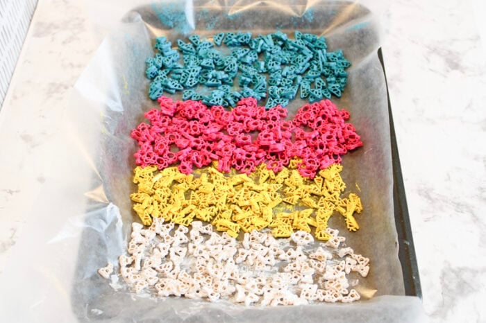 the white, yellow, pink, and blue painted bunny pasta is spread out on parchment paper in a baking pan.