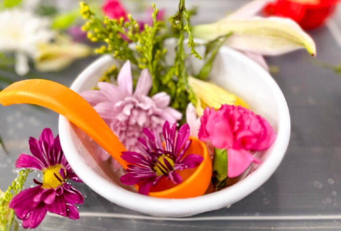a white bowl is full of colorful cut up flowers with a small orange ladle sitcking out.