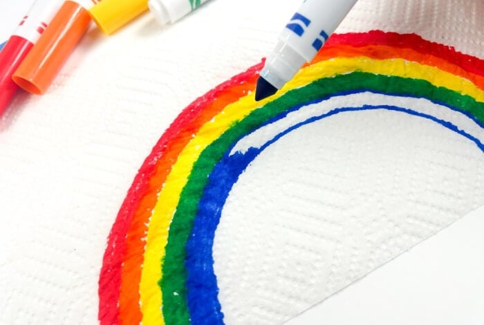 Magic Rainbow Science Experiment for Kids 16 someone is using a blue marker to draw a blue arc next to red, orange, yellow, & green arcs on a paper towel.