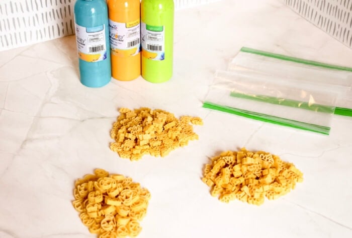 three equal piles of dried dinosaur pasta are on a counter next to paint and ziploc baggies.