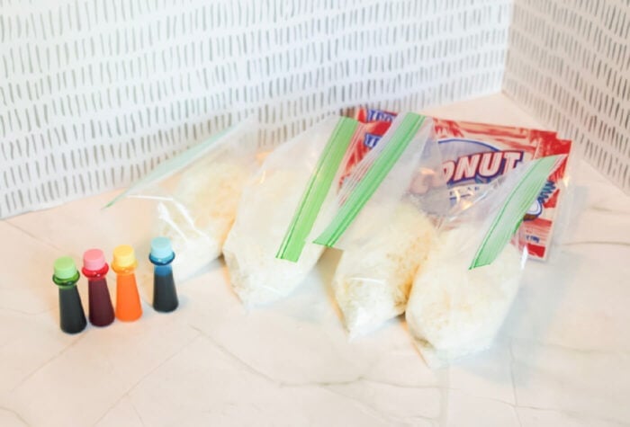 four ziploc bags of coconut flakes are sitting next to four bottles of food coloring.