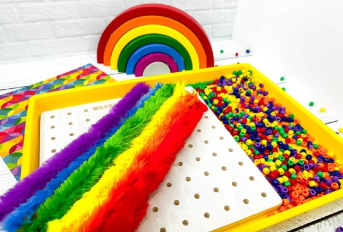 rainbow colored pipe cleaners, rainbow colored beans, and a small pegboard sitting inside a yellow tray.