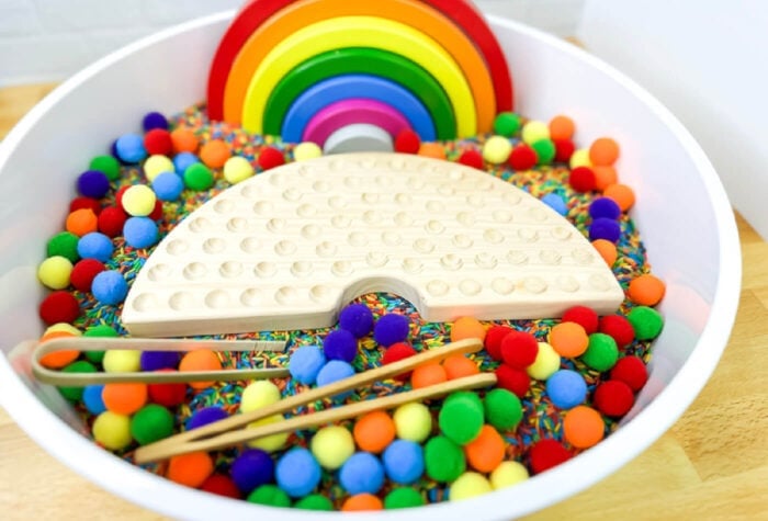 the rainbow sensory bin now has rainbow pom poms, tweezers, and a wooden pom pom holder in the shape of a rainbow.