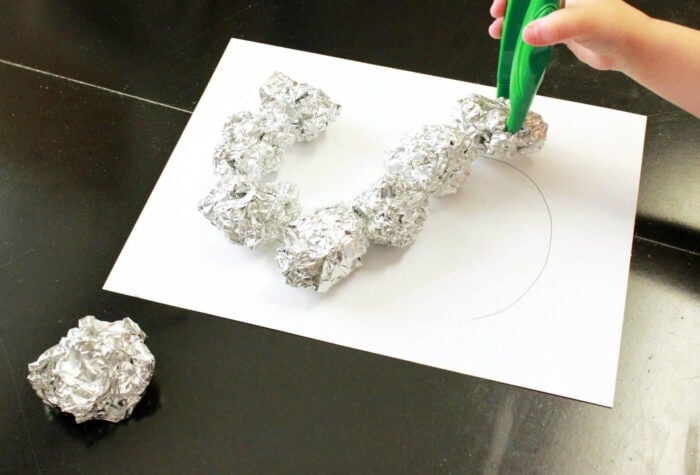 Exciting Outer Space Theme Lesson Plans for Preschool 12 a child using tweezers to place balls of aluminum foil on the outline of a letter "s".