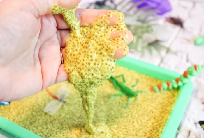 close up horizontal photo of a hand dripping with yellow chia seed slime. green sensory bin filled with chia seed slime and plastic bugs and insects in background