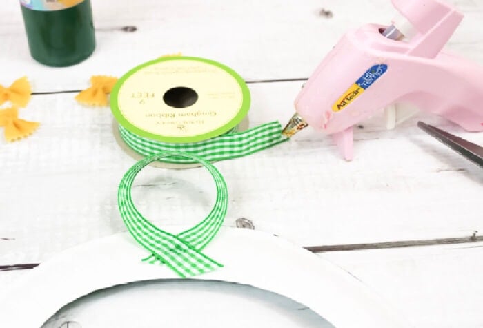 image of pink hot glue gun attaching green checked ribbon to the top of a paper plate wreath