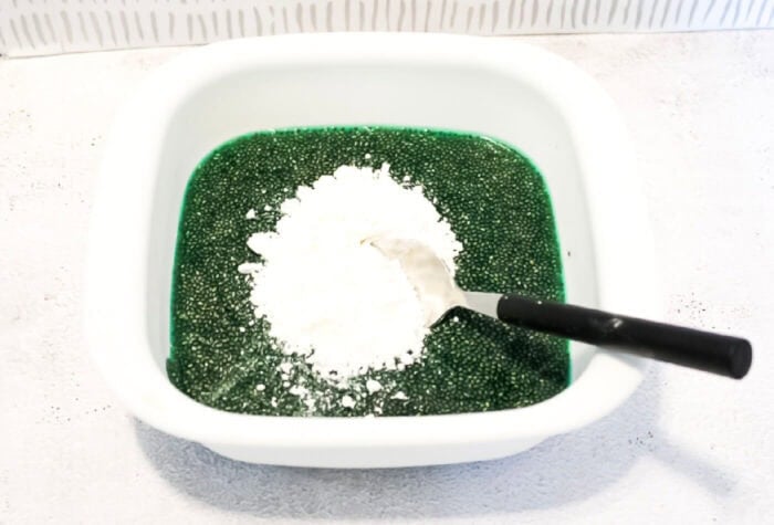 overhead image showing a spoon & a large scoop of cornstarch on top of a mixture of chia seeds, water, & green food coloring.