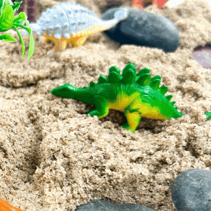 dinosaur theme activities for preschool and kindergarten featuring colorful toy dinosaurs exploring a sand sensory bin with rocks and greenery.